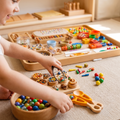 Toddler completely absorbed using wooden tongs to transfer rainbow beads between wooden bowls from Montessori Sensory Tray Kit on warm jute rug
