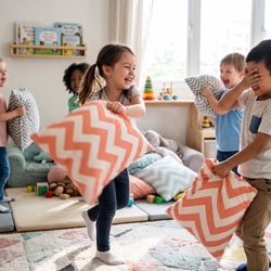Five children laughing and having an epic pillow fight with coral zigzag chevron cushions in a bright playroom