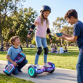 Girl in full protective gear riding Galaxy Chrome Hoverboard on sunny park path while brother controls via smartphone app and younger sibling watches excitedly