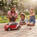 Three children in sunny garden playing with red Smart ForTwo RC car one steering with remote while younger children point and laugh at charging cable