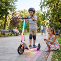 Boy in helmet and knee pads riding Rainbow LED Electric Scooter down sunlit park path with full rainbow LED strip glowing while sister smiles beside him