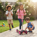 Girl in pink top scooting on pink 3-wheel kick scooter down sunny garden path while boy runs alongside clapping and toddler crouches to inspect wheels