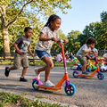 Girl with braids flying down sunny park path on Sunset Gradient Scooter laughing while boy sprints behind and younger sibling scoots in background